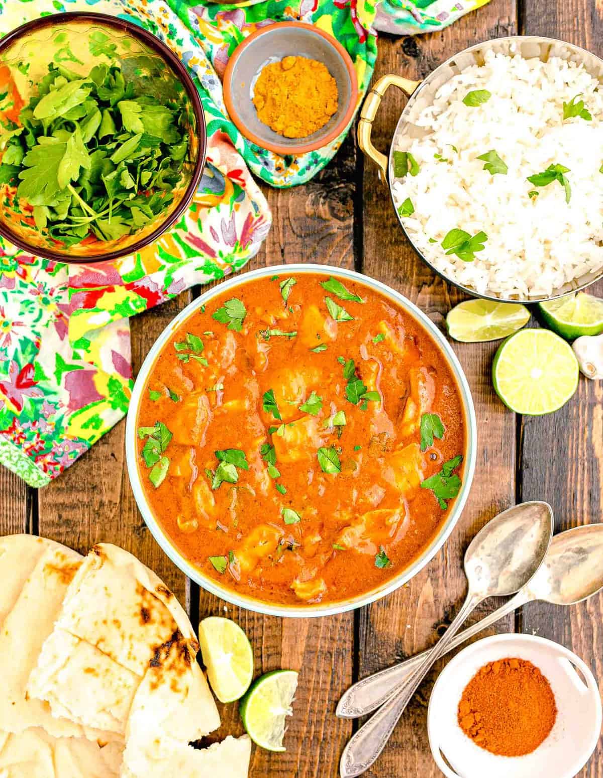 rice, cilantro and limes are placed around a bowl filled with butter chicken.