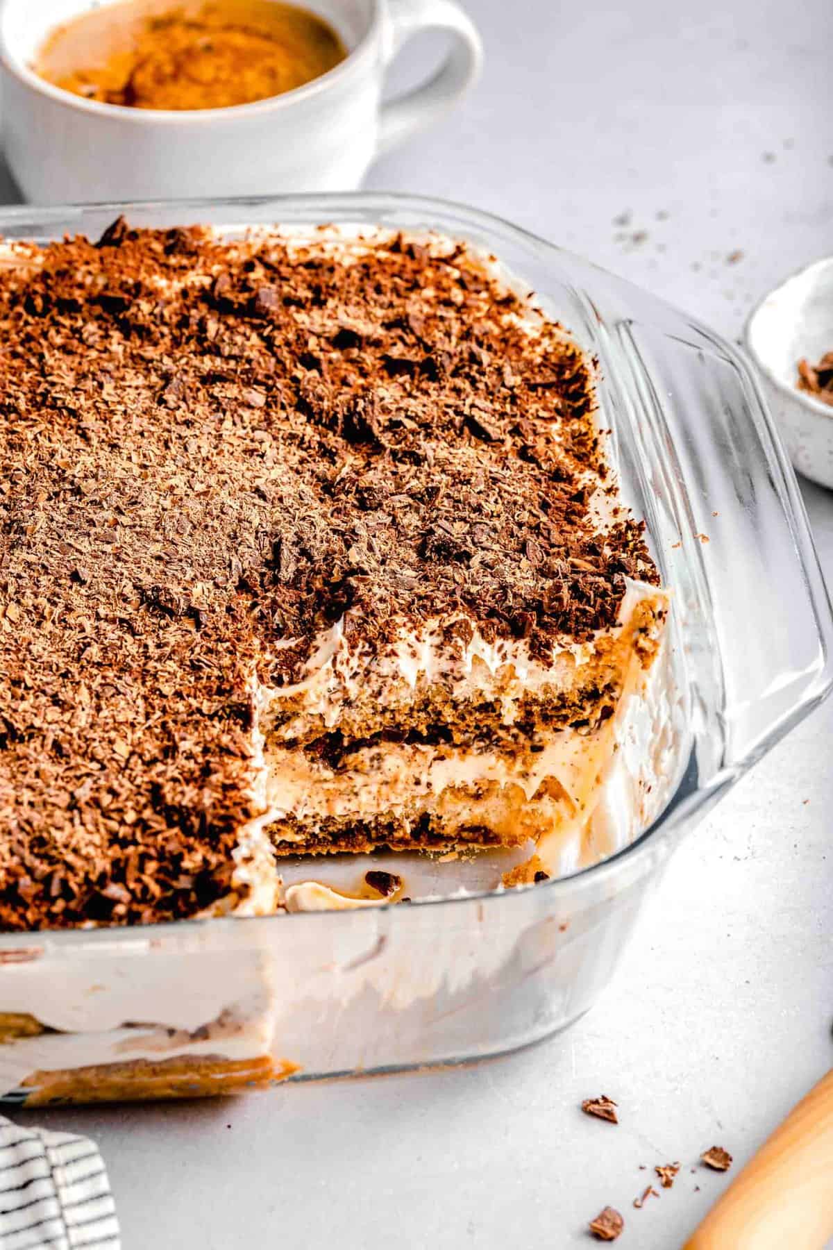 A baking pan of tiramisu with a slice taken out of it near a cup of espresso.