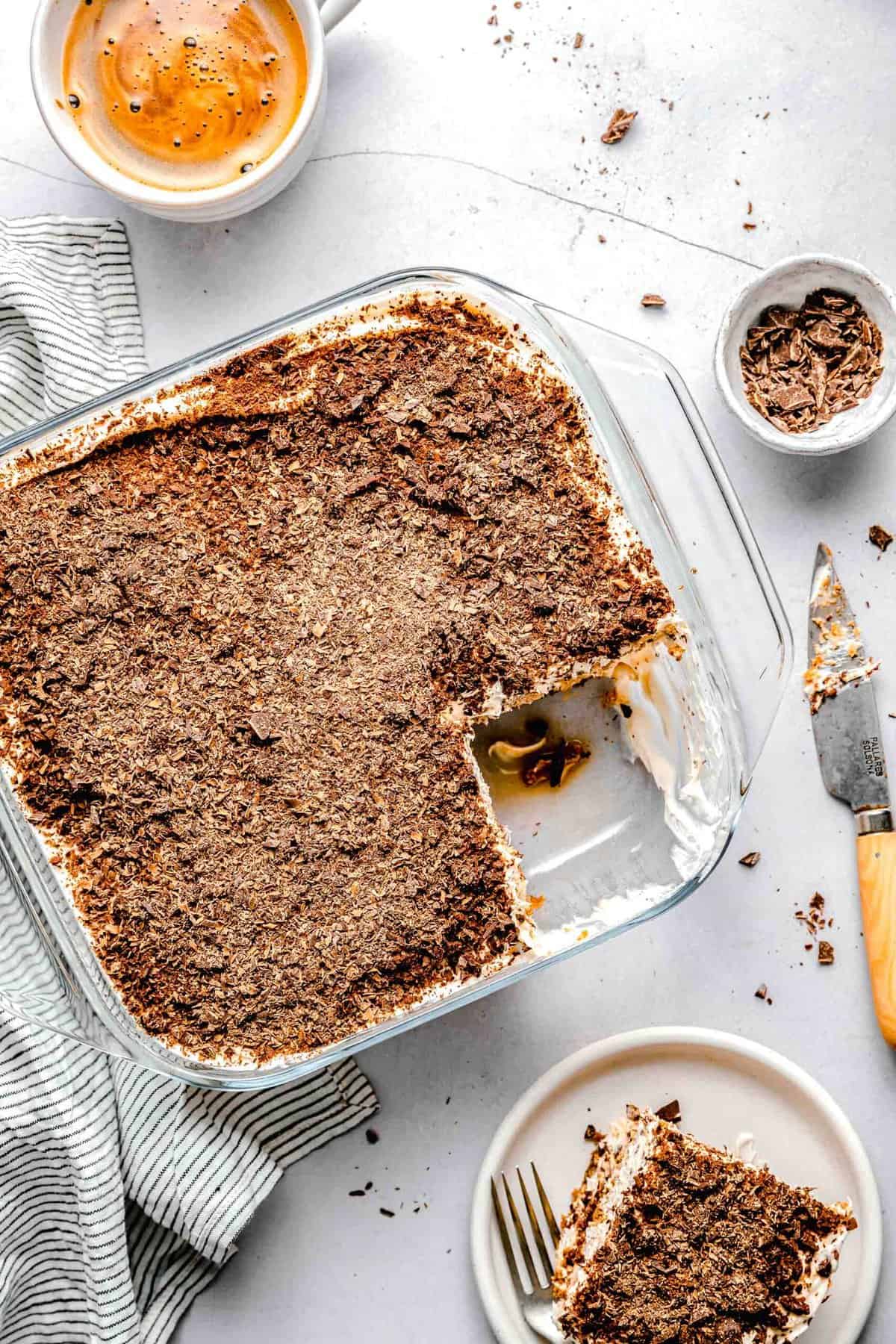 A baking pan of tiramisu with a slice taken out of it near a cup of espresso and a bowl of chopped chocolate.