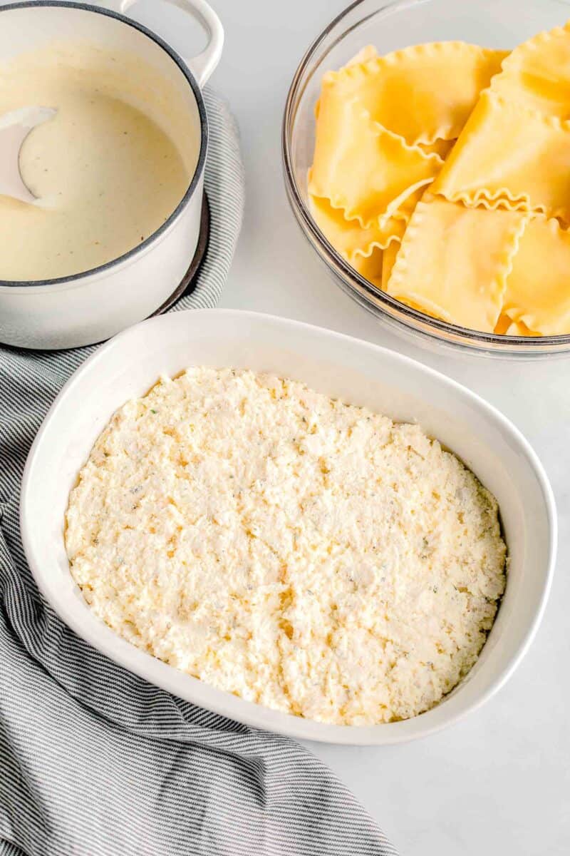 a white casserole dish is placed next to white sauce and lasagna noodles.