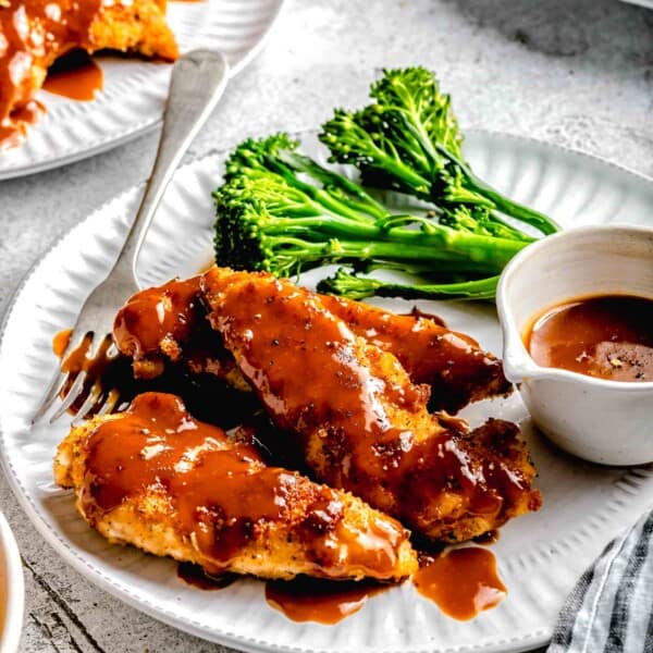 Side angle of bourbon chicken on a plate with broccoli, extra sauce, and a fork.