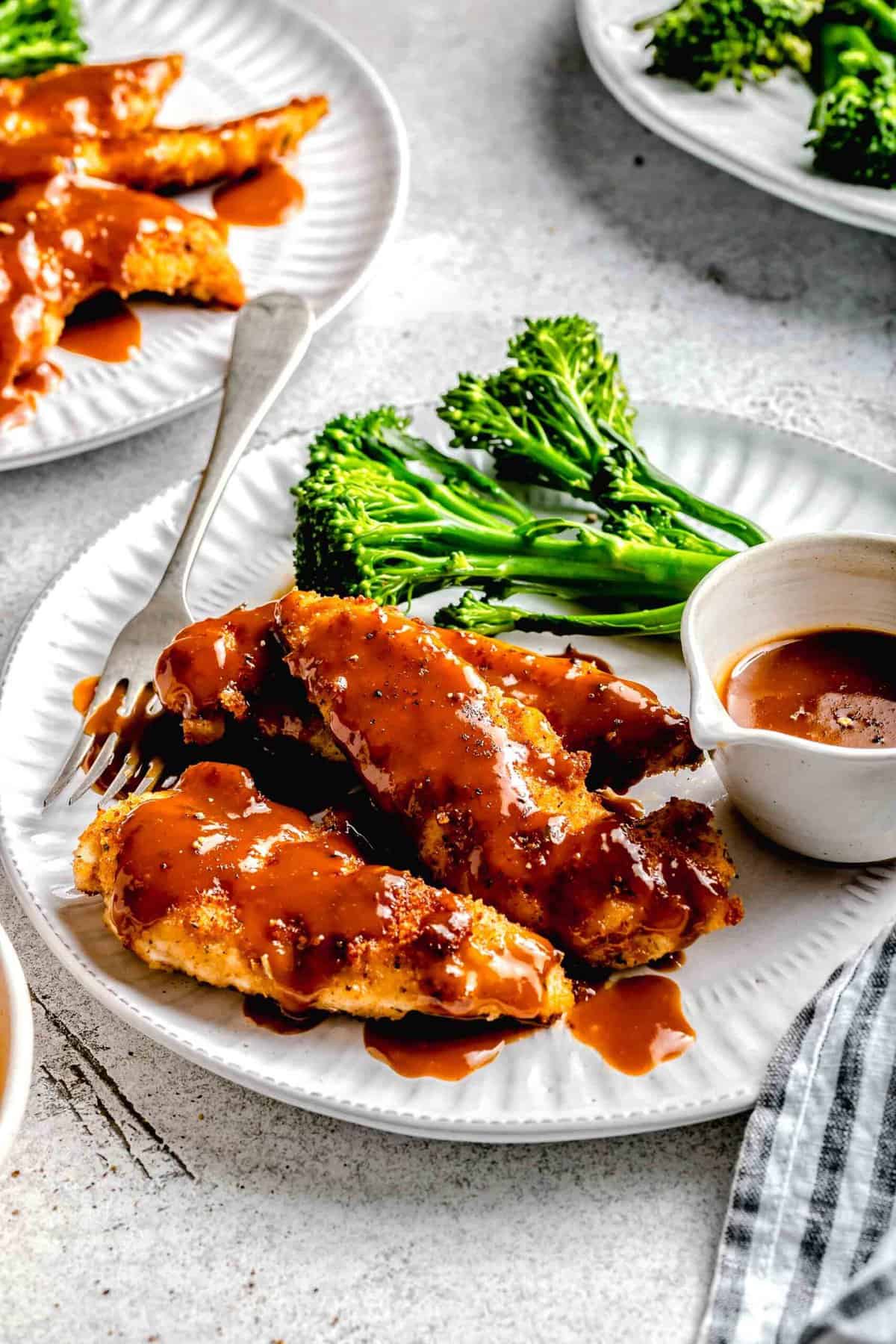 Side angle of bourbon chicken on a plate with broccoli, extra sauce, and a fork.