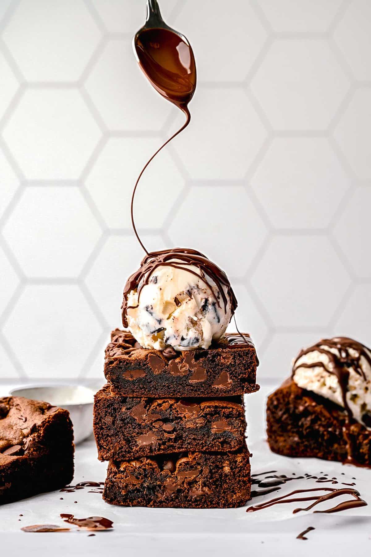 A stack of cake mix brownies with a scoop of ice cream on top and a spoon swirling chocolate sauce over the stack.
