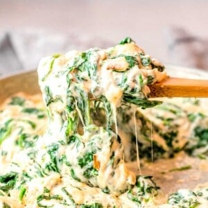 a wooden spoon is lifting creamed spinach from a skillet.