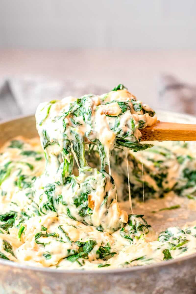 a wooden spoon is lifting creamed spinach from a skillet.