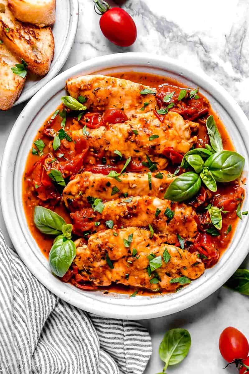 cooked chicken tenders lay on a bed of tomato herb sauce on a white plate with fresh basil