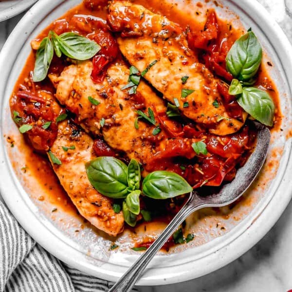 cooked chicken tenders lay on a bed of tomato herb sauce on a white plate with fresh basil and a metal spoon on the side