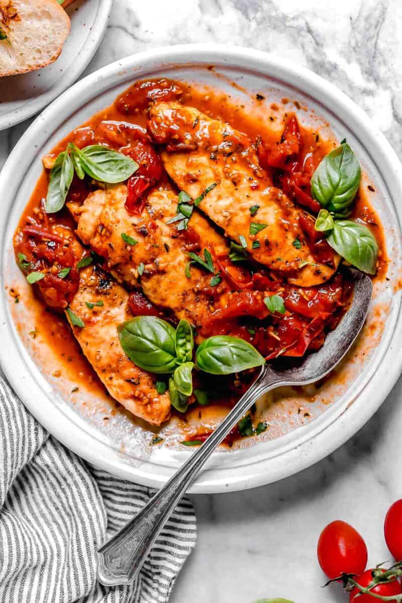 cooked chicken tenders lay on a bed of tomato herb sauce on a white plate with fresh basil and a metal spoon on the side