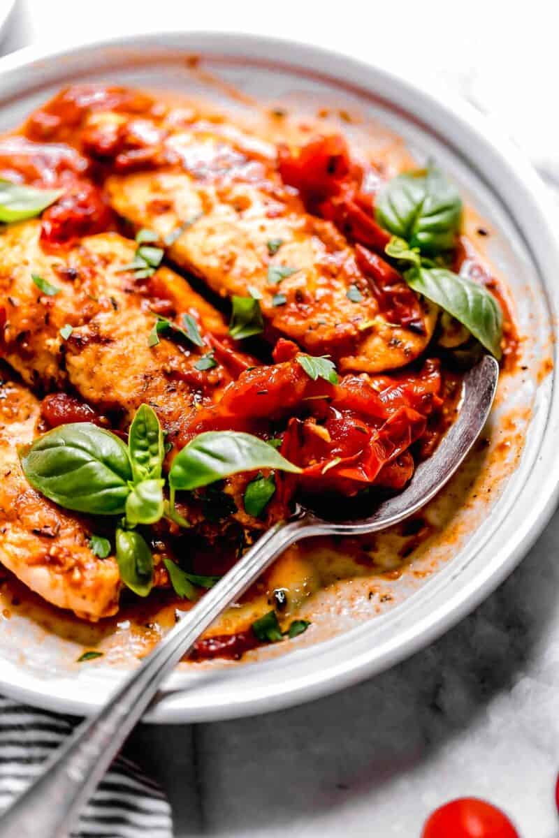 a metal spoon has tomato herb sauce next to cooked chicken tenders on a white plate