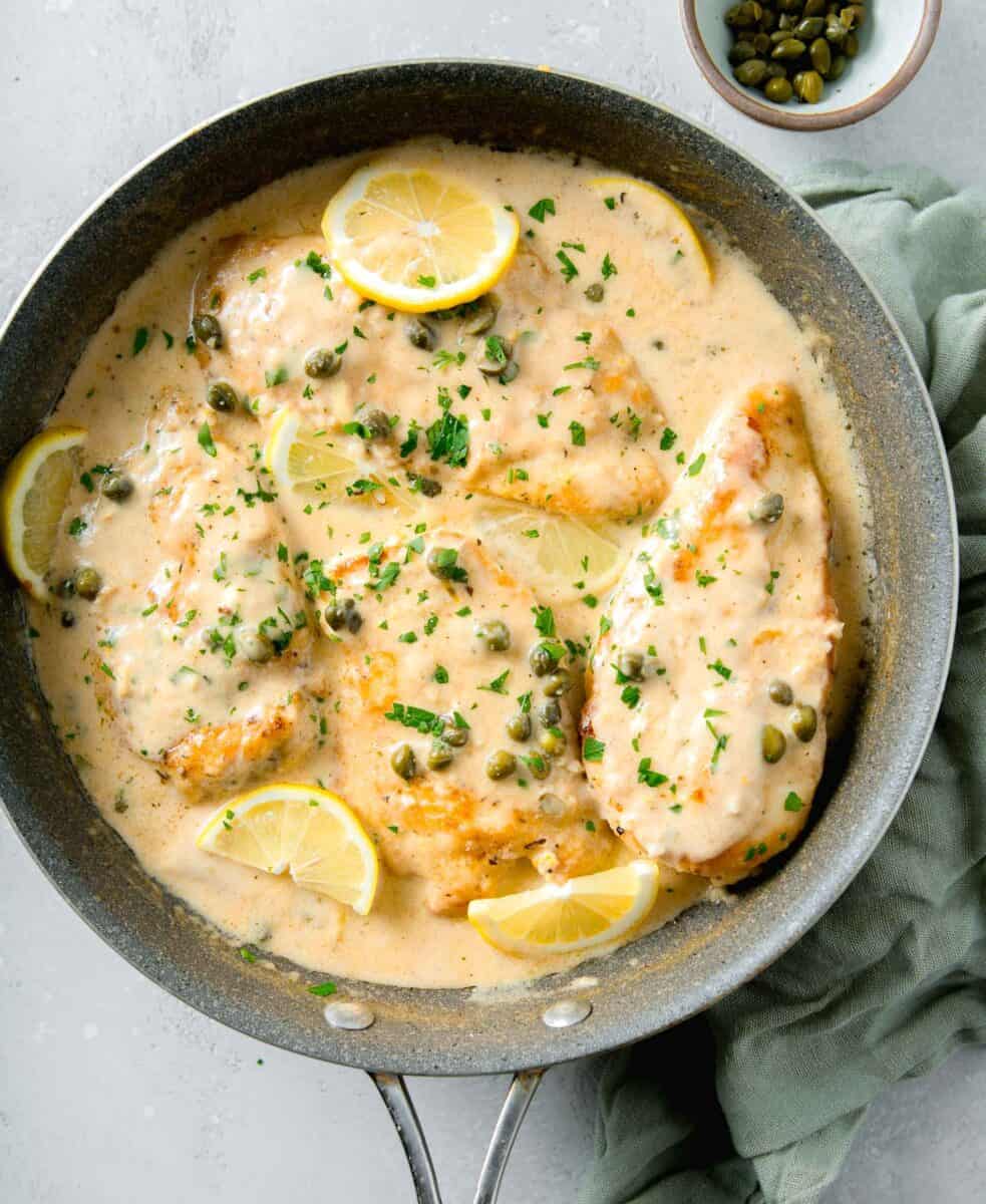 overhead image of four browned chicken breasts in a creamy lemon sauce with fresh parsley and capers on top finished with lemon slices.