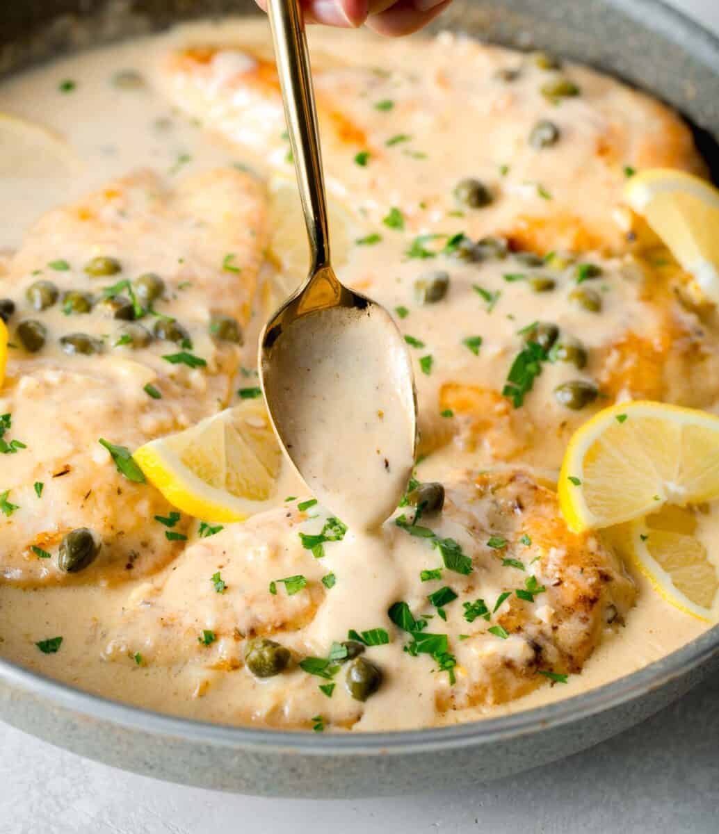 lemon cream sauce spooned on top of chicken breast in skillet next to lemon slices and fresh parsley and capers on top