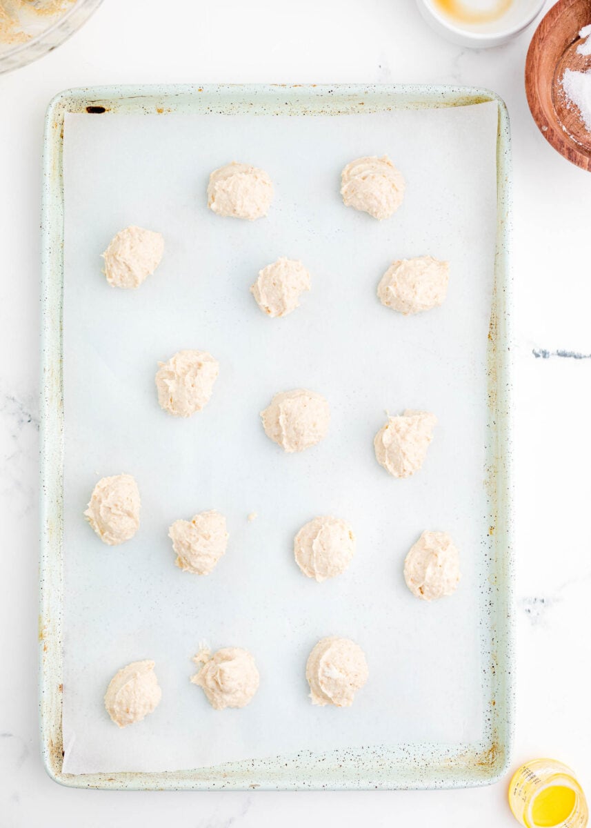 multiple white cream mixture balls are placed on a parchment paper lined baking sheet.