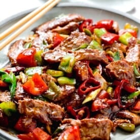 a large plate of hunan beef with wooden chopsticks sitting on the grey ceramic plate next to a light green towel