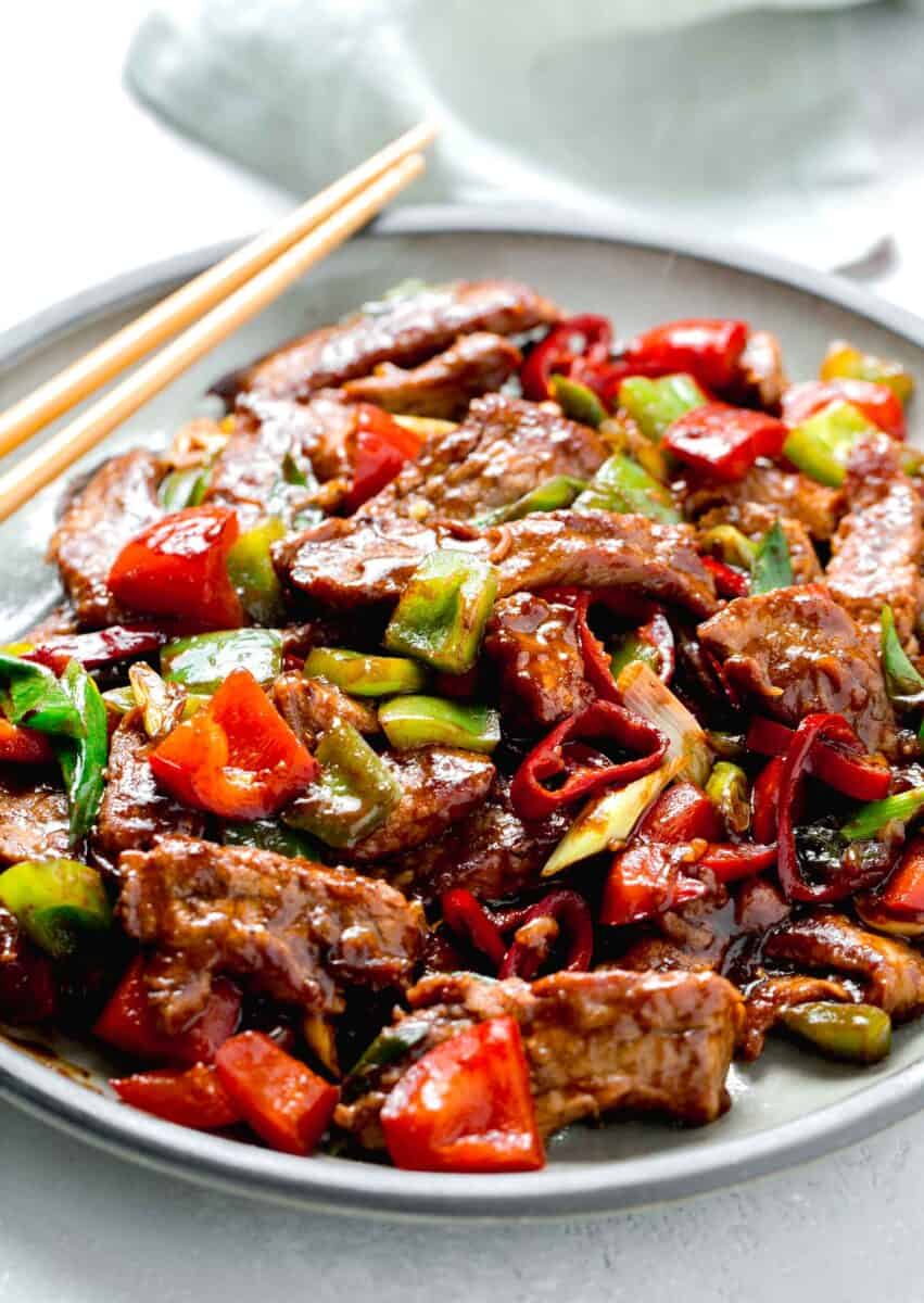 a large plate of hunan beef with wooden chopsticks sitting on the grey ceramic plate next to a light green towel