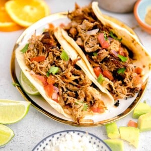 limes and oranges are placed all around a plate with two carnitas tacos on it.