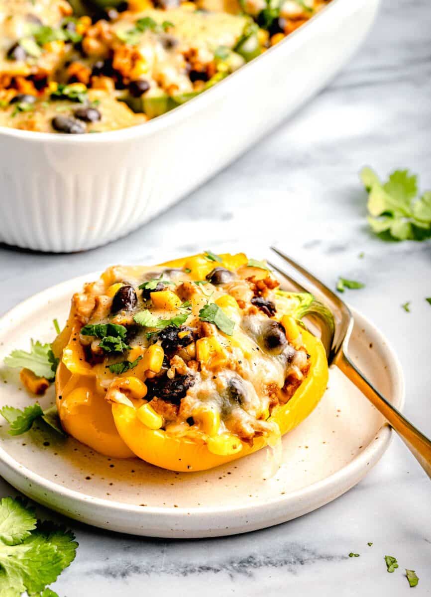 a single yellow stuffed pepper on a white speckled plate with a gold fork next to the larger white baking dish