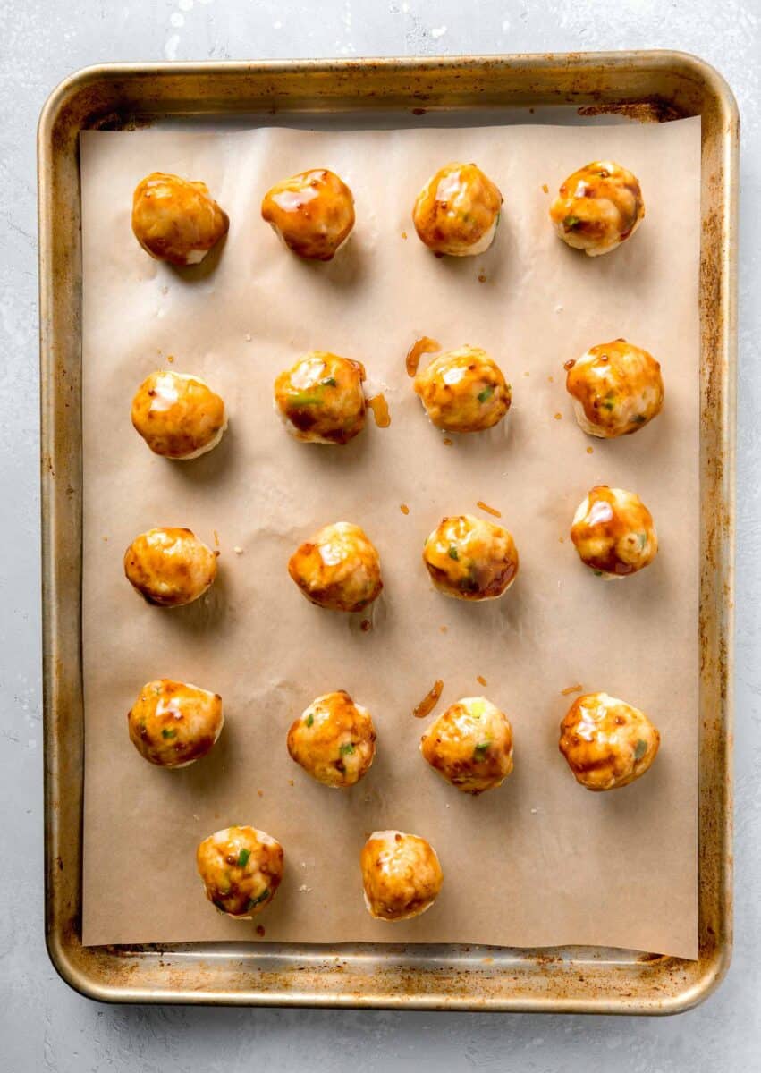 teriyaki turkey meatballs formed into meatballs with teriyaki sauce brushed on. the rolled meatballs are on top of parchment paper on a rimmed baking sheet