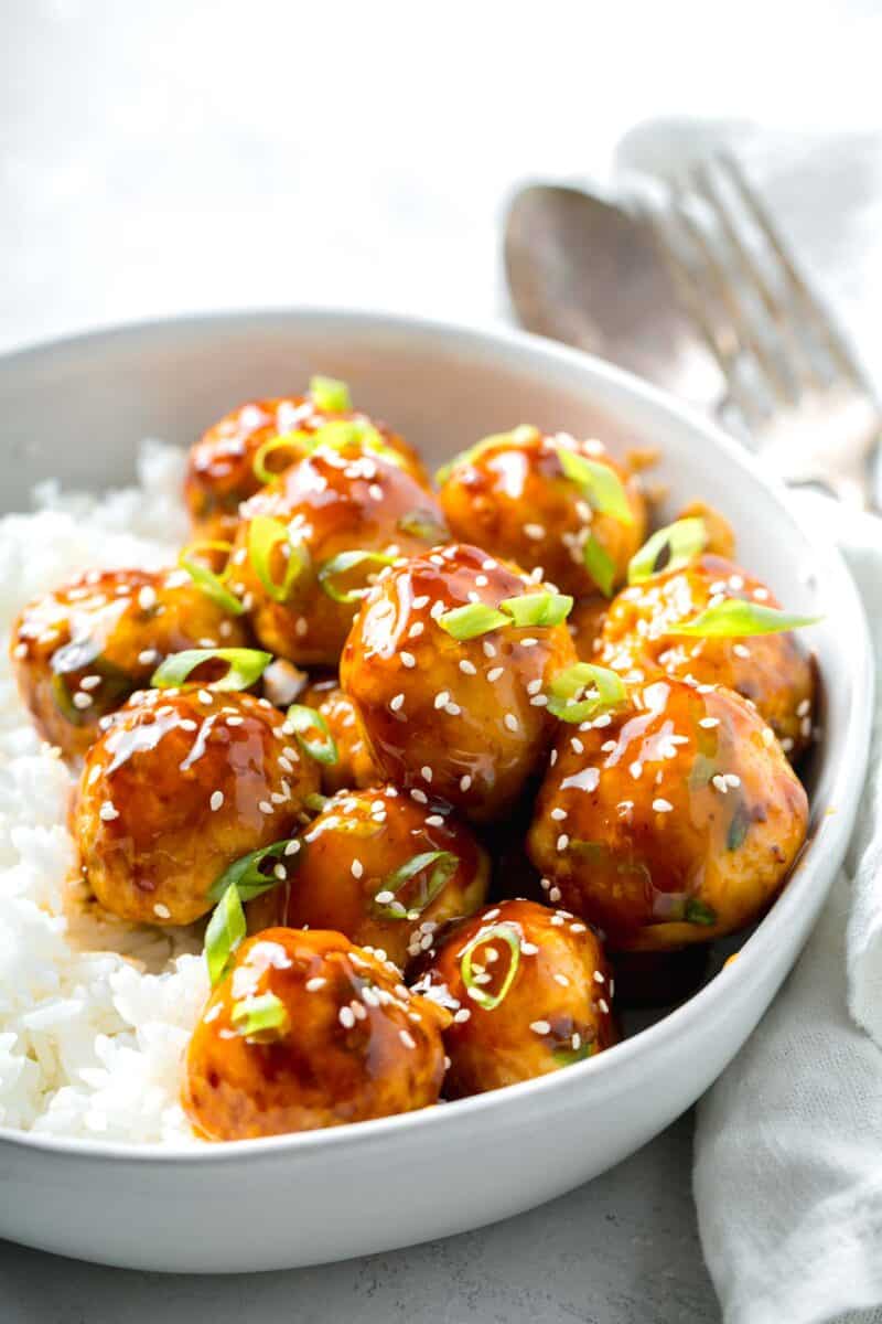 teriyaki chicken meatballs glazed in teriyaki sauce with fresh scallions and sesame seeds in a bowl with white rice next to a linen napkin