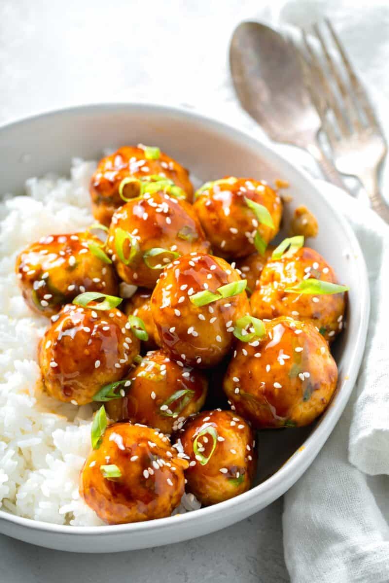 teriyaki chicken meatballs glazed in teriyaki sauce with fresh scallions and sesame seeds in a bowl with white rice next to a linen napkin