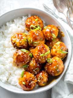 overhead image of teriyaki chicken meatballs glazed in teriyaki sauce with fresh scallions and sesame seeds in a bowl with white rice next to a linen napkin and a fork and spoon