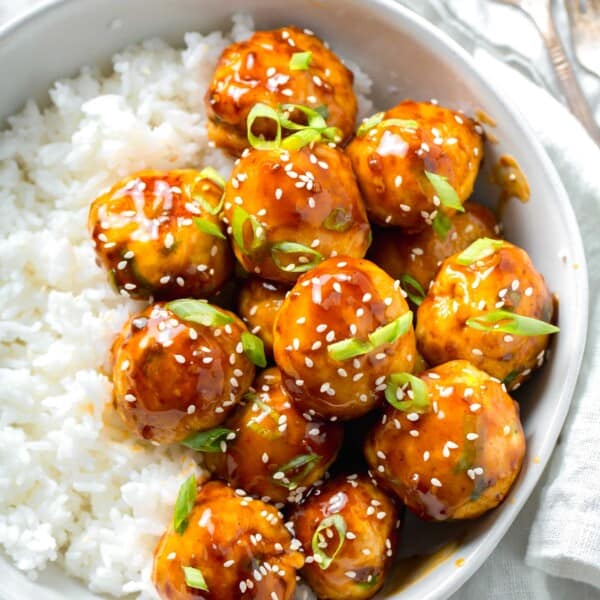 overhead image of teriyaki chicken meatballs glazed in teriyaki sauce with fresh scallions and sesame seeds in a bowl with white rice next to a linen napkin and a fork and spoon