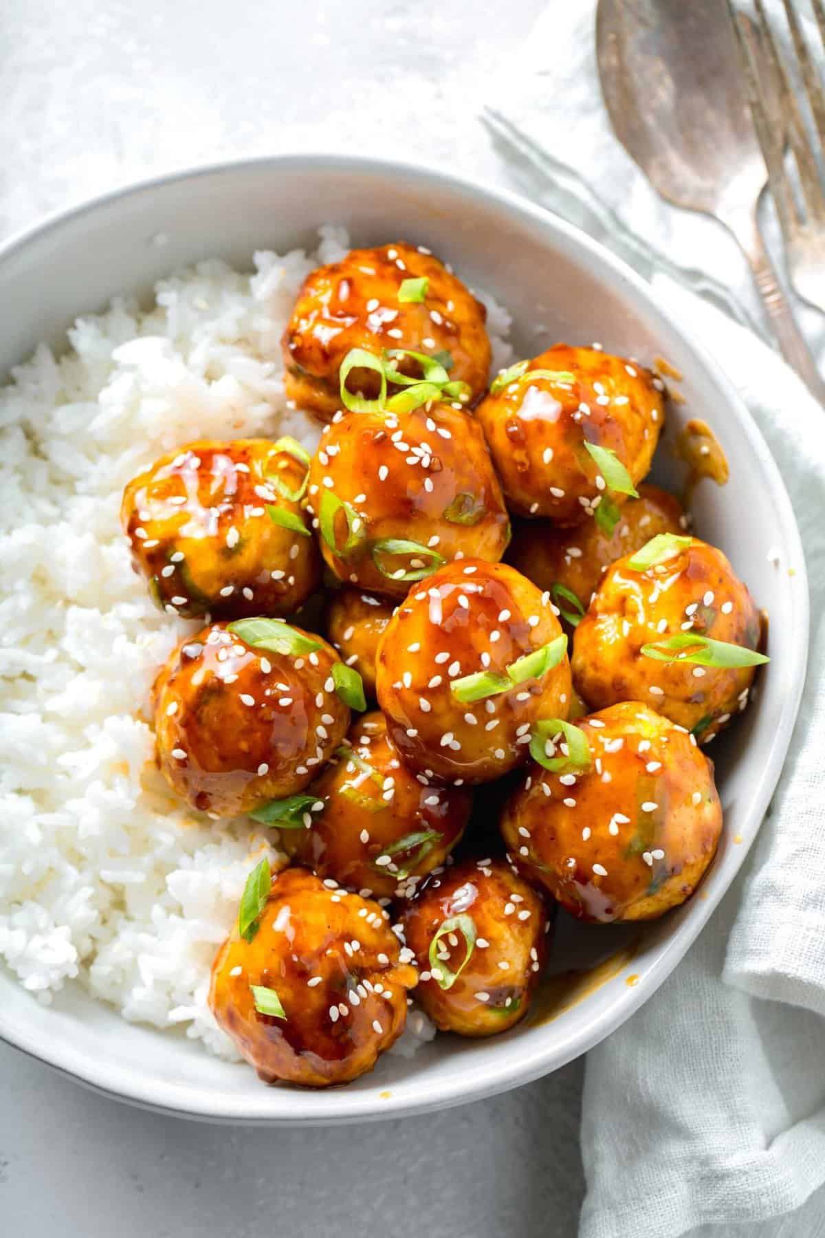 overhead image of teriyaki chicken meatballs glazed in teriyaki sauce with fresh scallions and sesame seeds in a bowl with white rice next to a linen napkin and a fork and spoon