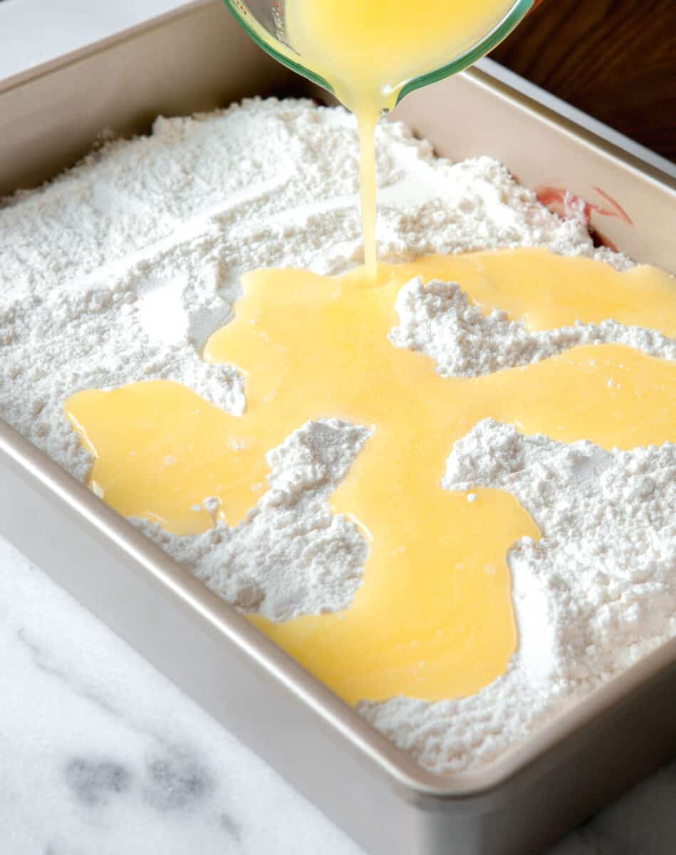butter being poured on top of the white cake mix in the metal baking pan