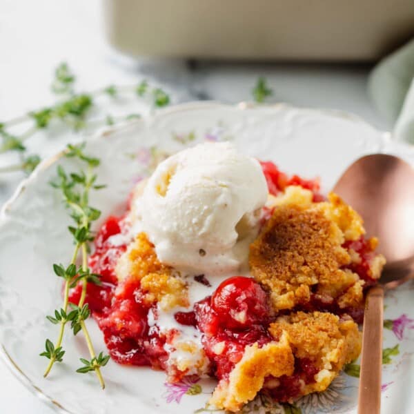 cherry dump cake on a floral dessert plate with a scoop of vanilla ice cream on top and sprigs of thyme