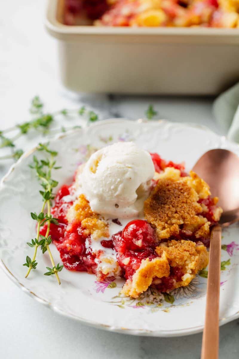 cherry dump cake on a floral dessert plate with a scoop of vanilla ice cream on top and sprigs of thyme