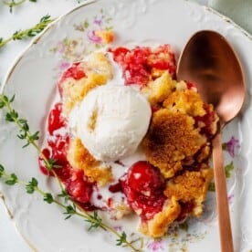 overhead image of cherry dump cake a la mode on a floral dessert plate next to a copper spoon and sprigs of thyme