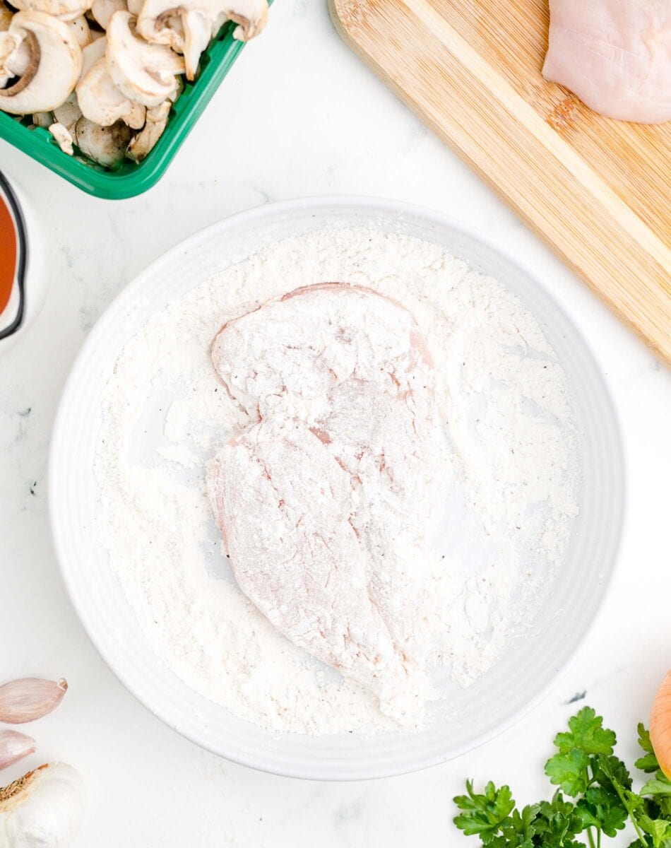 a flour coated chicken breast is in a shallow white bowl.