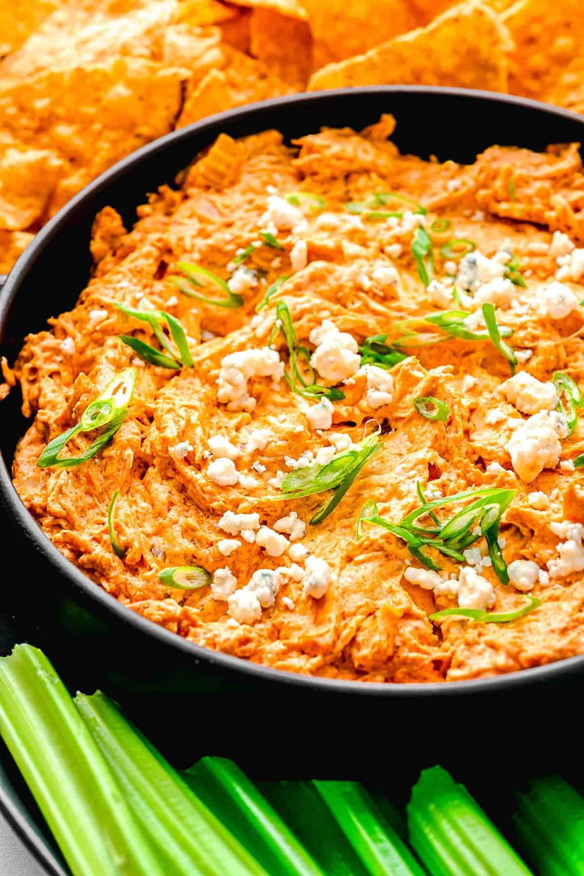 Closeup of buffalo chicken dip in a serving bowl near celery sticks and tortilla chips.