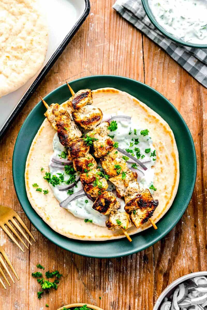 greek chicken souvlaki skewers on top of pita with tzatziki sauce and fresh parsley on a green plate