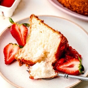 A slice of strawberry angel food cake on a plate with fresh strawberries and a fork taking a bite out of it.