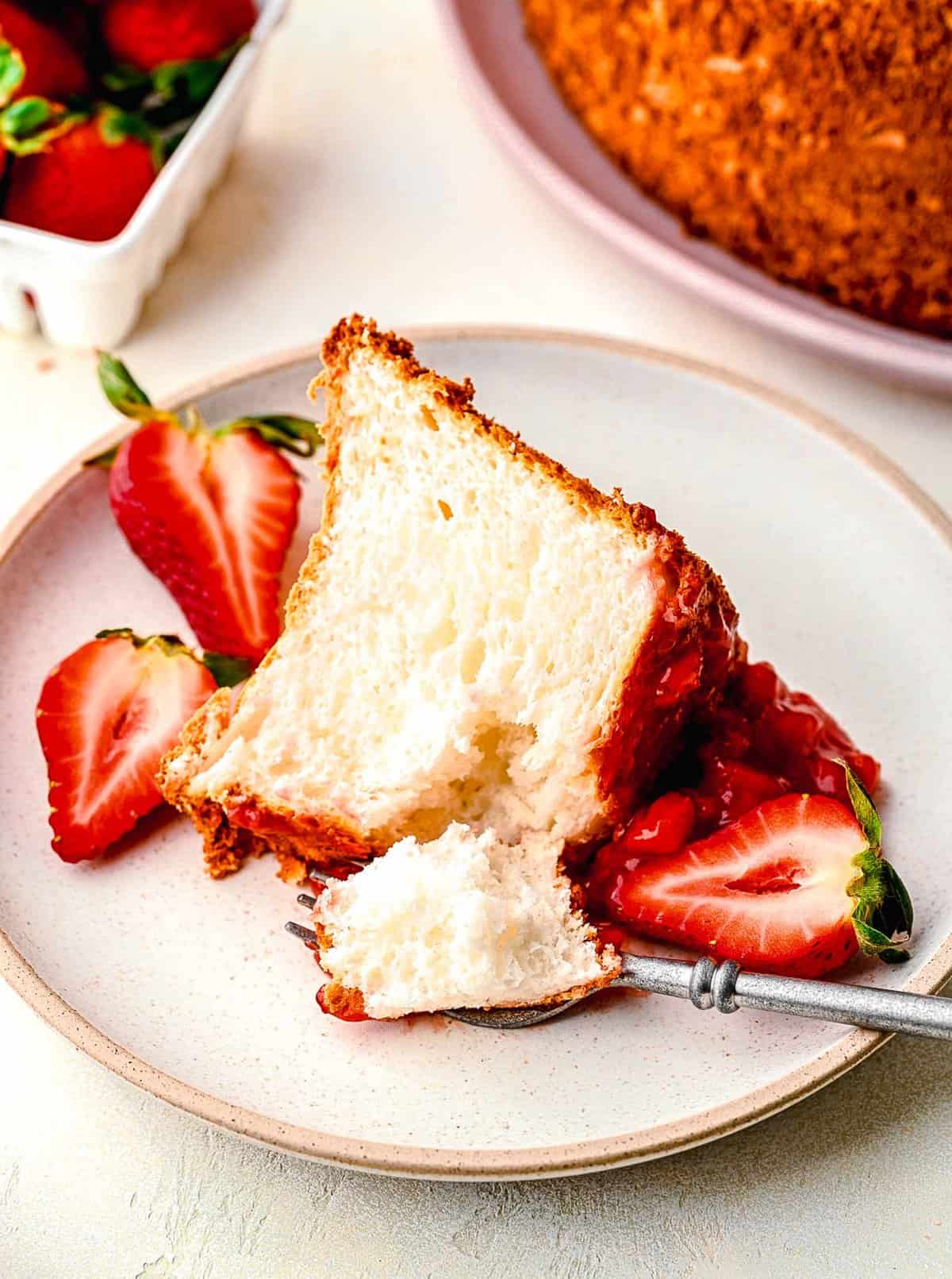 A slice of strawberry angel food cake on a plate with fresh strawberries and a fork taking a bite out of it.