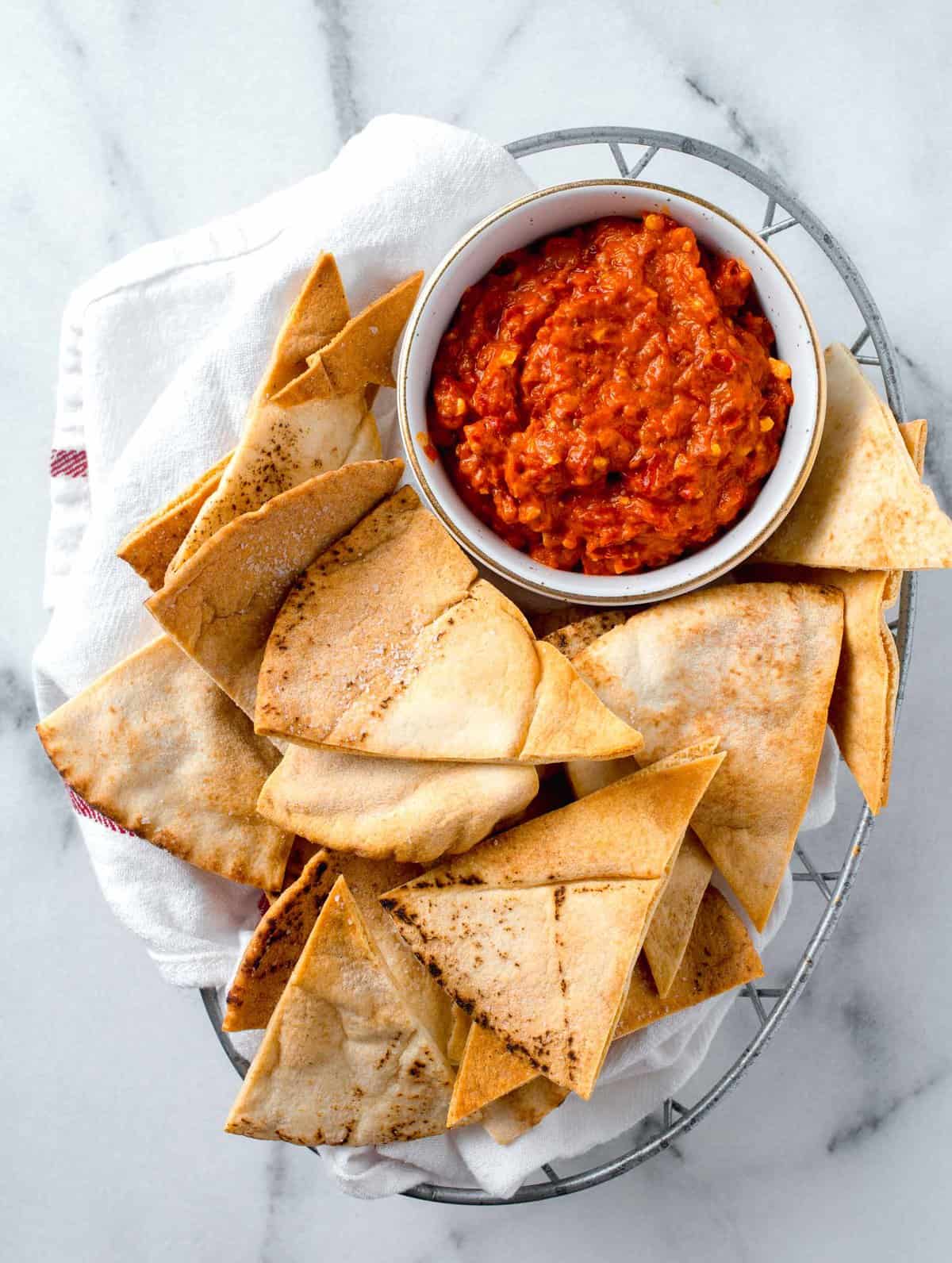 air fried pita bread in a metal basket lined with a linen towel next to a white bowl of red pepper dip