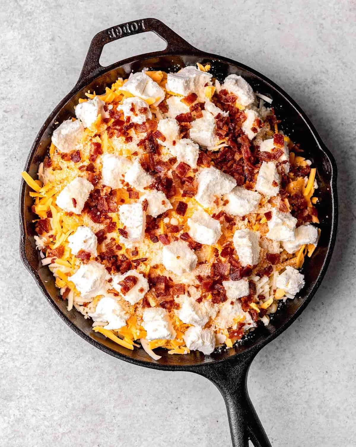 all the ingredients for corn dip in a cast iron skillet. cubes of cream cheese and bacon bits can be seen on top