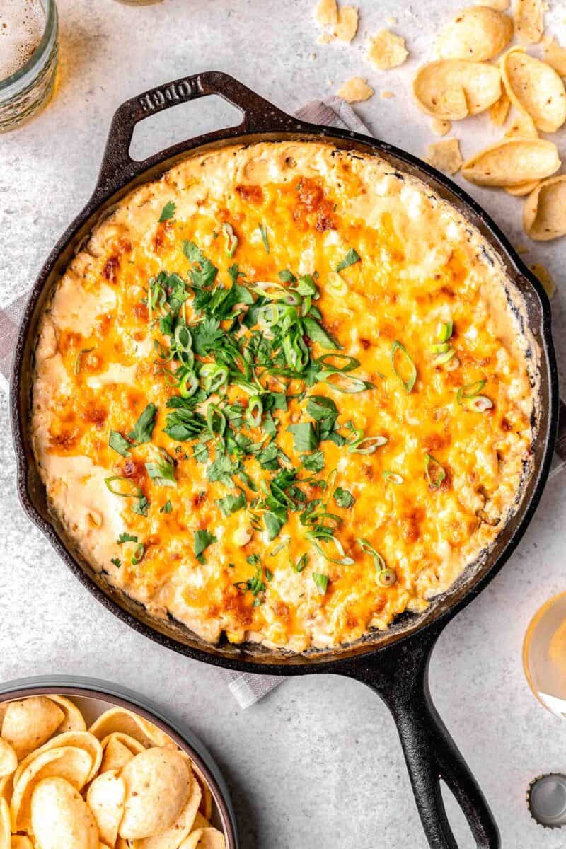 freshly baked corn dip in a cast iron skillet with fresh cilantro and green onions sprinkled on top next to tortilla chips and beer