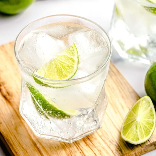 A gin and tonic on a cutting board near a slice of lime. Another gin and tonic is in the background.