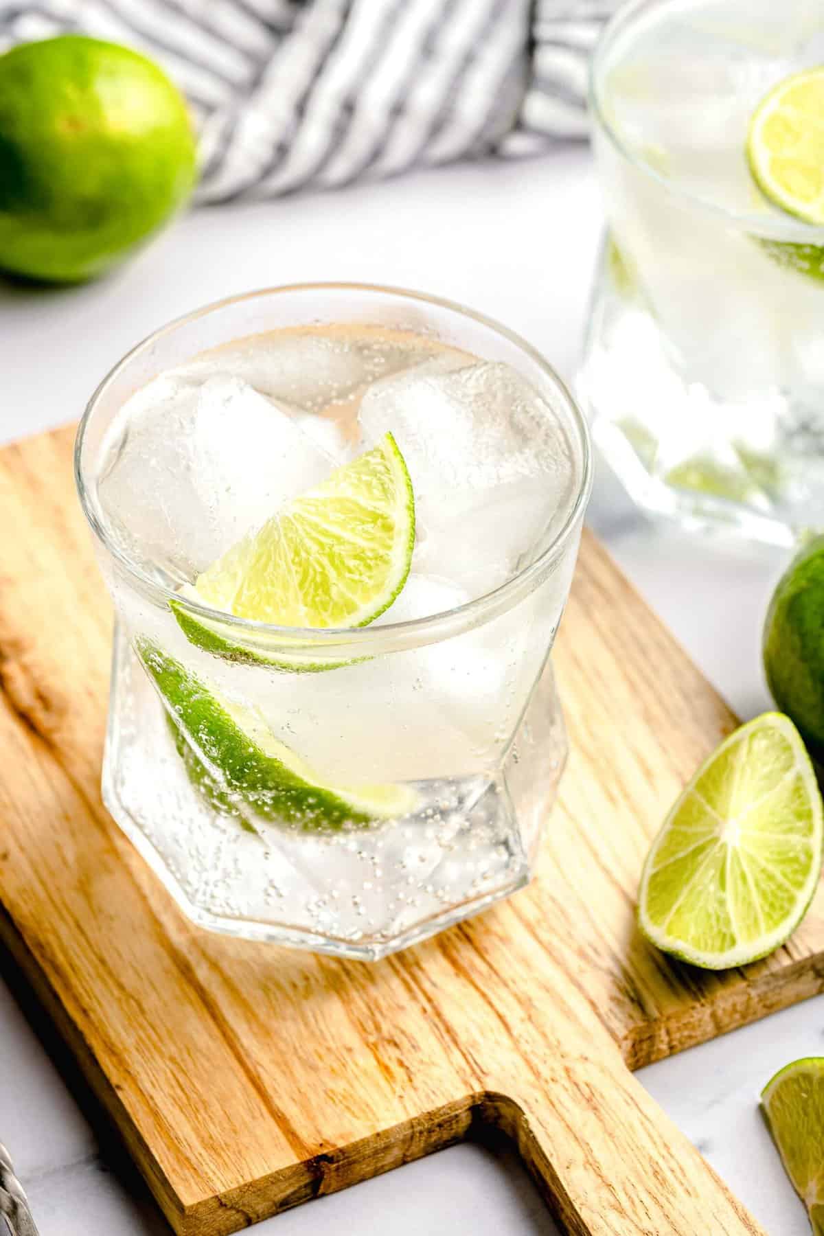 A gin and tonic on a cutting board near a slice of lime. Another gin and tonic is in the background.