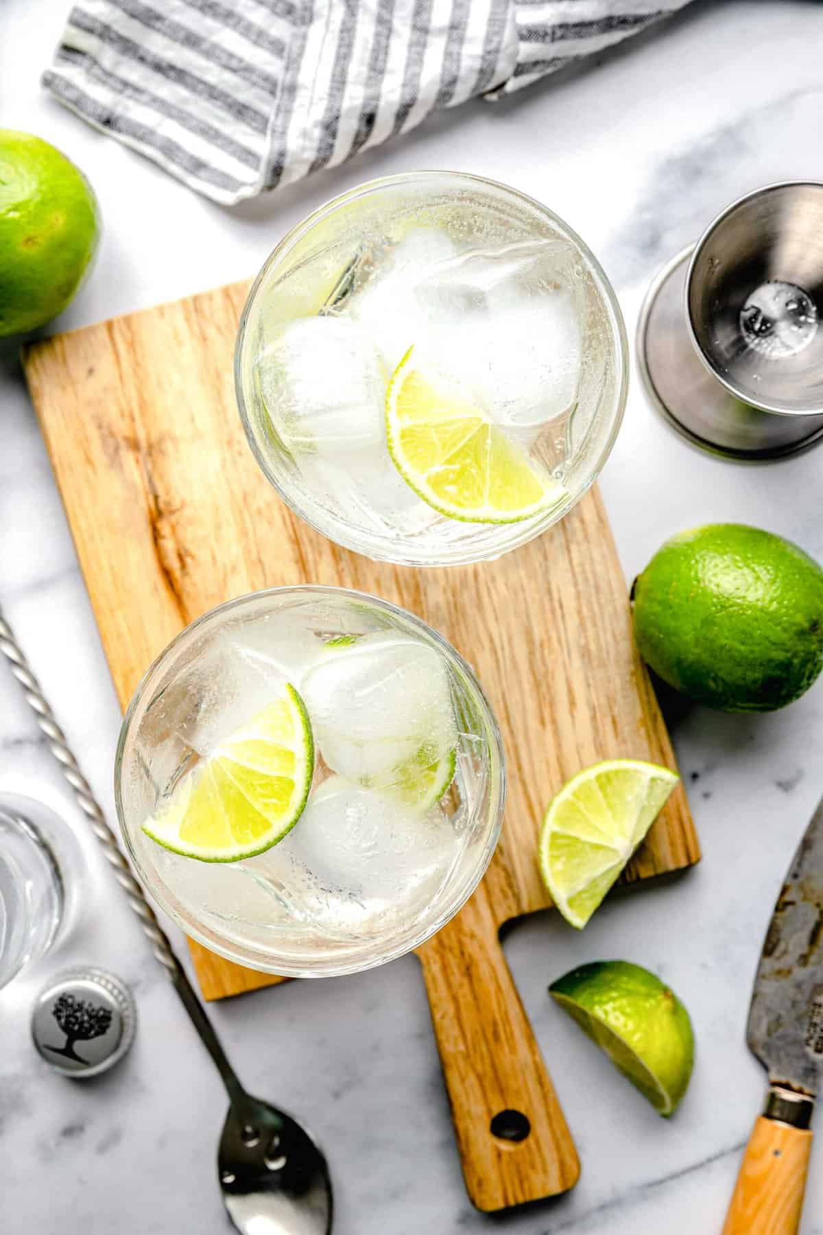 Overhead of 2 gin and tonics on a cutting board with a slice of lime surrounded by cocktail mixing accouterments.