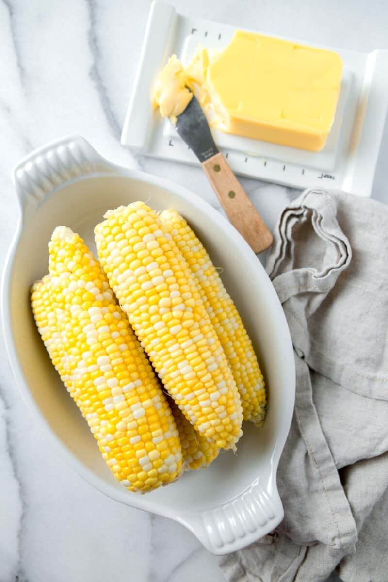 cooked corn on the cob in a casserole dish next to a linen towel and butter