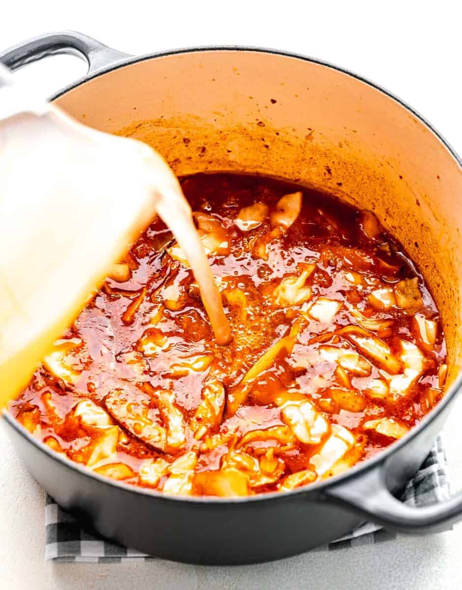 Adding chicken broth to a mixture of gochujang paste, kimchi juice, and sautéed mushrooms, kimchi, and garlic.