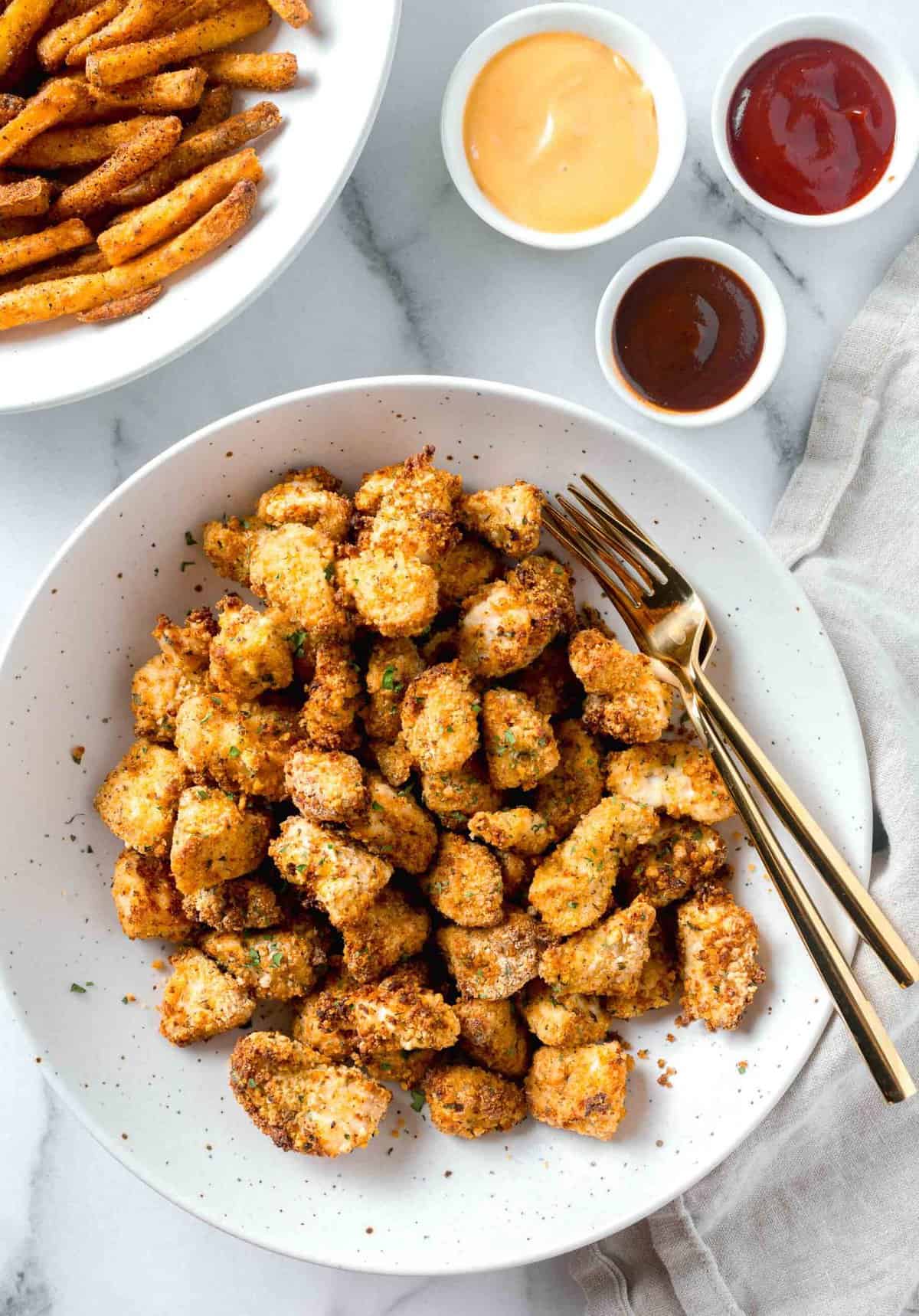 air fryer chicken nuggets in a large speckled white bowl with two gold forks next to a bowl of fries and various dipping sauces