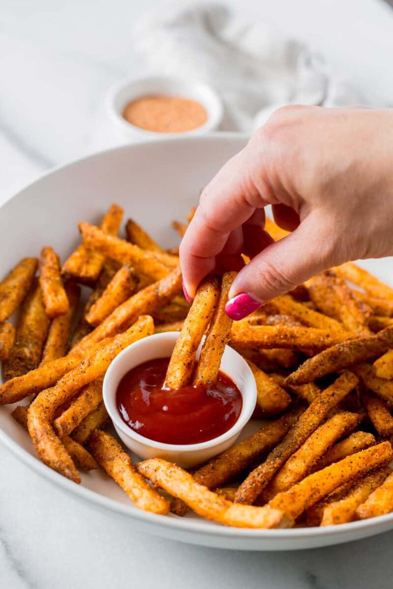 a hand is holding two fries and dipping into ketchup on a plate of cajun fries