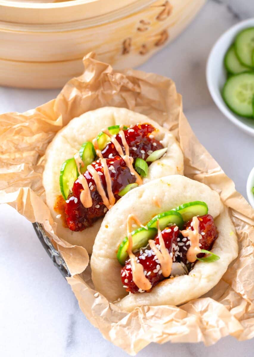 chicken bao buns on brown parchment paper next to a steamer and cucumbers