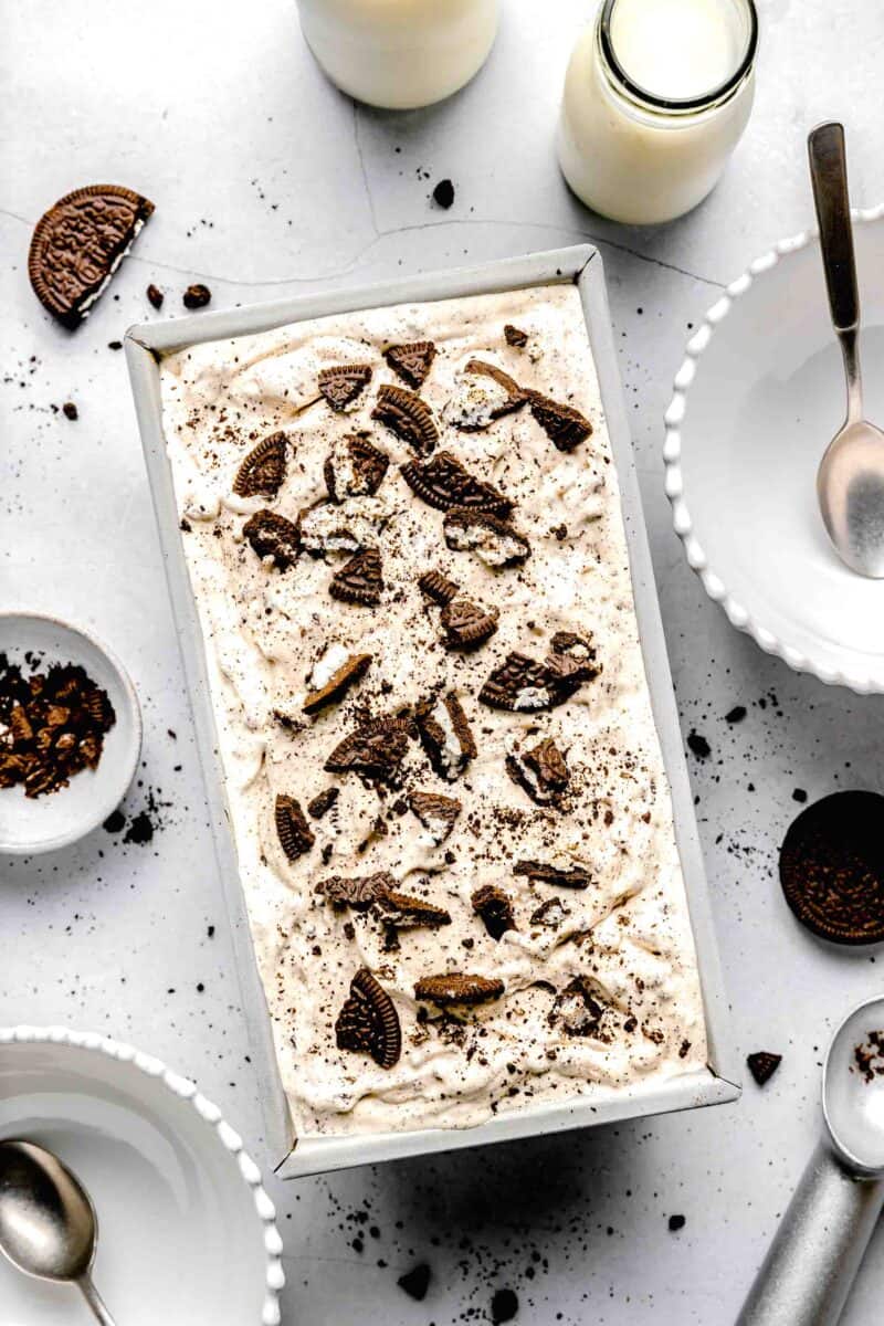 cookies and cream ice cream in a straight loaf pan with crushed cookies surrounding the pan and milk and empty bowls
