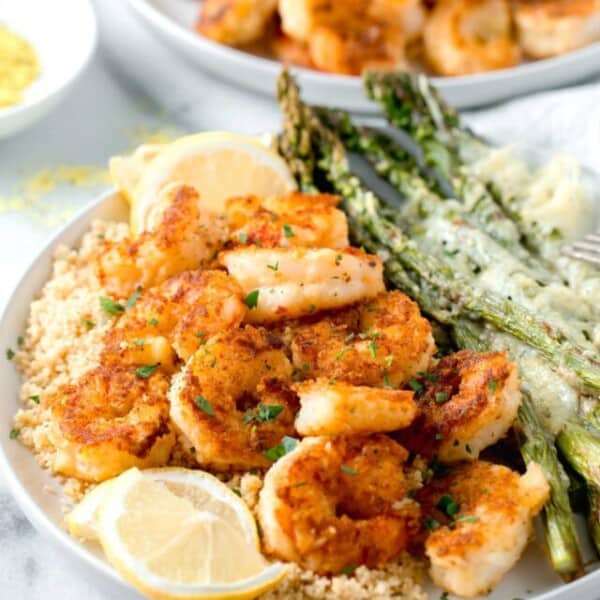 lemon pepper shrimp on top of couscous and next to parmesan crusted asparagus on a white plate with fresh lemon slices