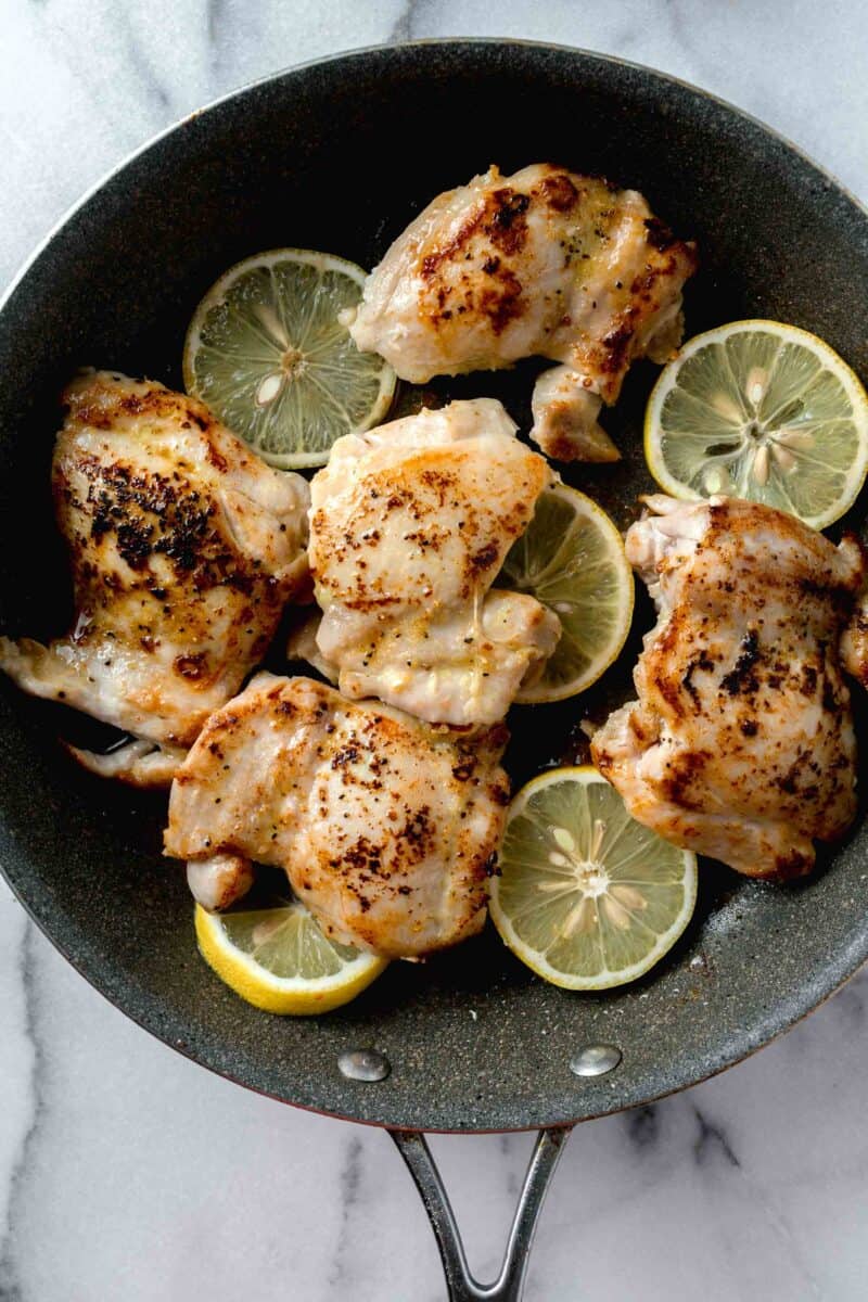 seared chicken thighs with fresh lemon slices in a grey nonstick skillet