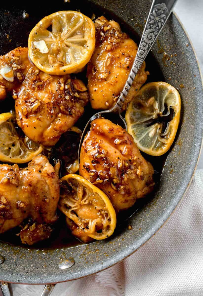 honey garlic lemon pepper chicken thigh in a grey skillet under a metal spoon next to fresh lemon slices